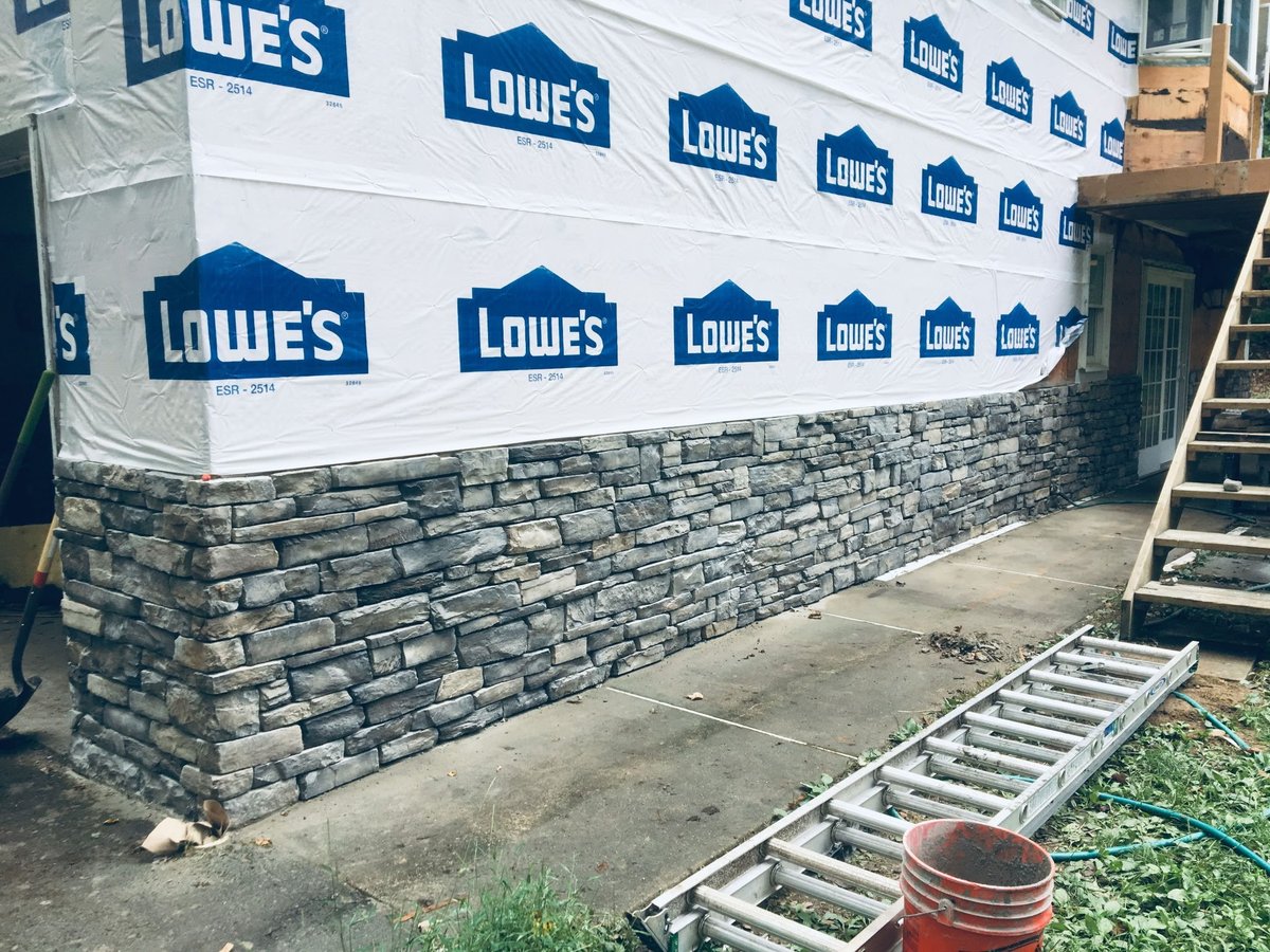 Lowes branded blue and white protective wrapping covering a stone brick wall under construction, with metal ladder and red bucket visible in foreground