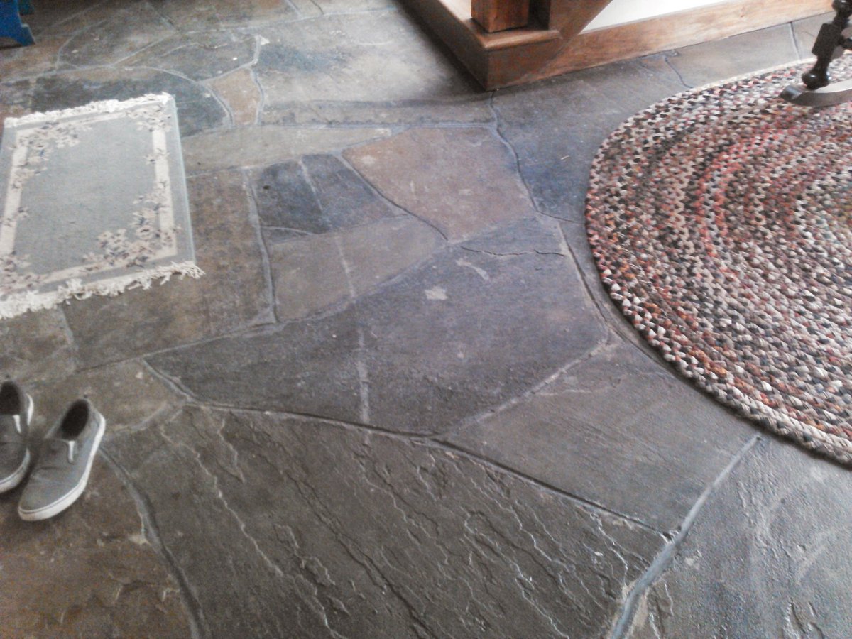 Overhead view of a dark slate floor with geometric patterns, a round woven mat, white vessel, and wooden furniture