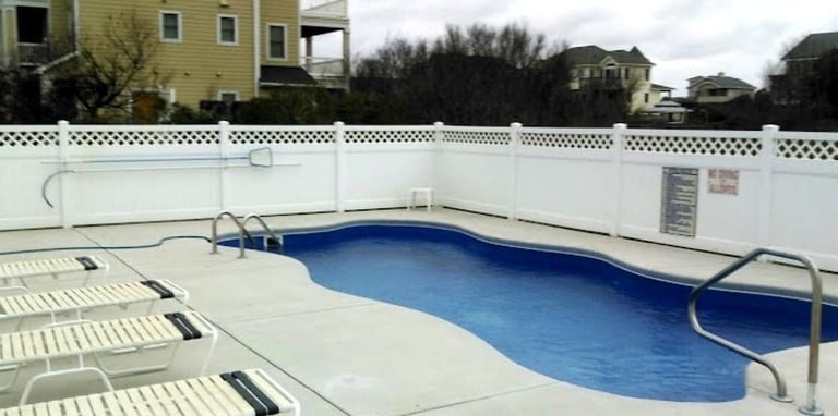 Empty residential swimming pool with blue water, white deck, metal railings, and white picket fence bordering a suburban neighborhood