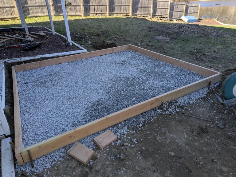 Raised garden bed filled with gray gravel inside a fenced yard with green lawn and white support posts
