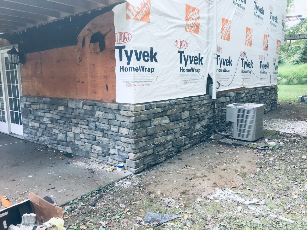 Construction exterior wrapped in Tyvek HomeWrap material with stone veneer base and HVAC unit visible on the ground