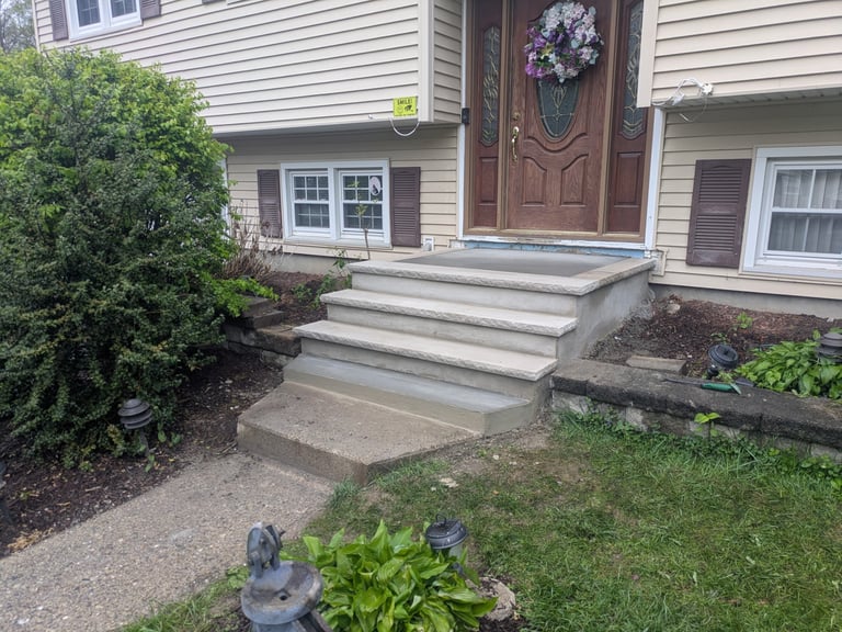 Two-story house with gray siding, brown front door, white steps leading to entrance, decorative hedges, flower beds, and lawn in foreground.