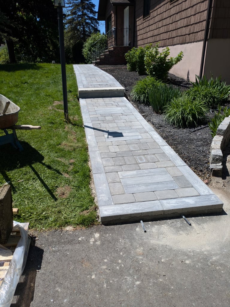 Newly installed light gray paver pathway leading to a house entrance, bordered by green lawn and landscaped plantings