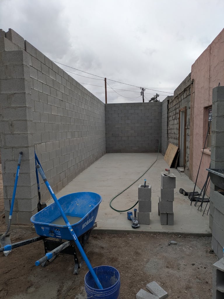 Construction site with concrete courtyard, blue wheelbarrow, cinder blocks, and cement mixer under gray overcast sky