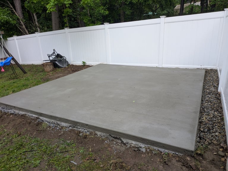 Newly poured concrete pad in backyard with white vinyl fence, trees, and gravel border
