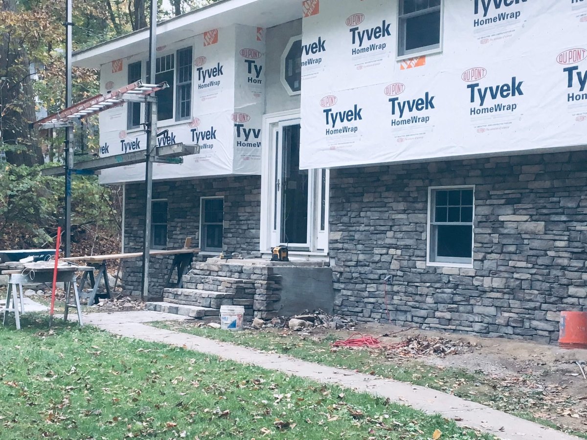 Two-story house under construction with Tyvek wrap on upper level and stone veneer cladding on lower level, surrounded by landscaping